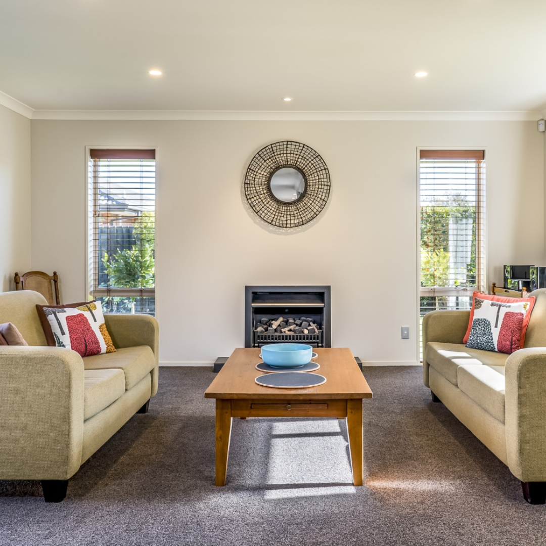 home builder’s services Living room with two beige sofas, a wooden coffee table, a round wall mirror, and a small fireplace. Large windows on either side allow natural light to illuminate the space.